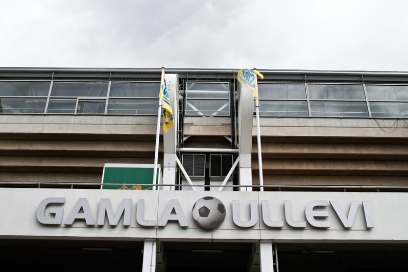 En bild som visar en del av en byggnad med texten "GAMLA ULLEVI" i stora grå bokstäver. Mellan orden finns en större symbol föreställande en fotboll. Två gulblåa flaggor syns bredvid skylten, och ovanför syns fönsterrader och betongstrukturer.