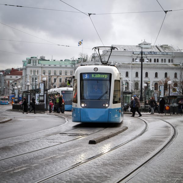 En spårvagn med destination "Kålltorp" är i förgrunden till höger, och bakom den passerar en annan spårvagn. I bakgrunden syns folk som väntar vid en hållplats och på andra sidan gatan ser man byggnader. Himmelen är grå och molnig.
