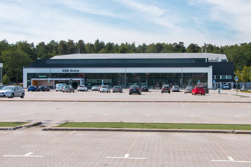 En stor, modern byggnad med texten "ABB Arena" syns i bilden, omgiven av en parkeringsplats med flera parkerade bilar. Grönskande träd bildar en bakgrund och himlen är ljusblå med moln. Marken består av grå marksten med vita parkeringslinjer.