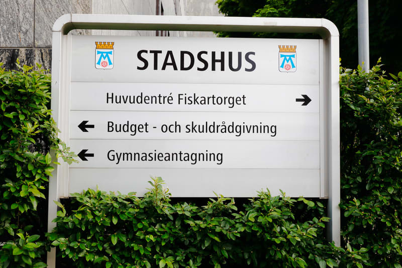 En vit skylt med texten STADSHUS och två vapensköldar omgiven av gröna buskar. Skylten pekar mot Huvudentré Fiskartorget, Budget- och skuldrådgivning, samt Gymnasieantagning med pilar.