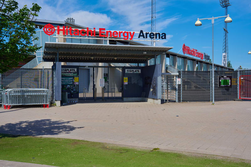 En fasadvy av en arena med skylt "Hitachi Energy Arena" mot en blå himmel med moln. En entré med grindar och staket är synlig, och ett område med gräs och träd i förgrunden.