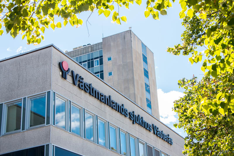 En bild av en byggnad med texten "Västmanlands sjukhus Västerås" och en logotyp. Ljusa gröna lövverksgrenar är synliga i förgrunden uppe och till höger, och himlen är blå med några moln. Byggnaden har många fönster.