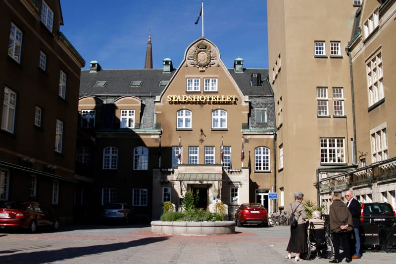 En fasadbild av "Stadshotellet" med två bilar parkerade vid stenläggning. Några människor står vid en svarta bil nära hotellet, och i förgrunden syns skuggor på marken. En mörk bil med registreringsnummer "OGV 263" står till vänster, delvis i skugga.