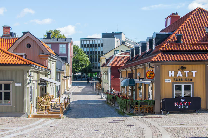 En gata med gatsten i en stadskärna, flankerad av byggnader i olika färger, främst gula och beige, med röda takpannor. Träd bidrar med grönska i bakgrunden. Två restauranger med uteserveringar syns på sidorna.