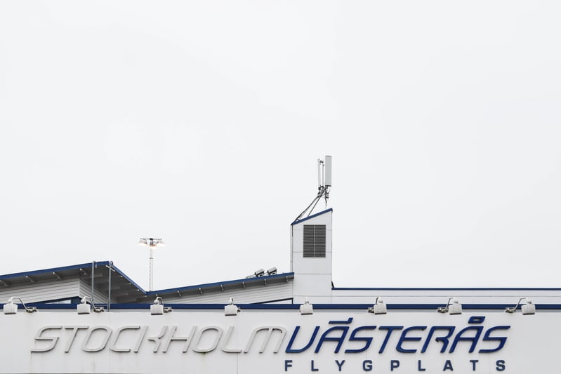 En bild som visar en del av en byggnad med skyltning för 'STOCKHOLM VÄSTERÅS FLYGPLATS'. På taket finns belysningsarmaturer, antenner och en ventilationslucka, med en ljus, molnig himmel i bakgrunden.