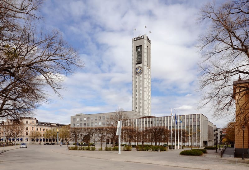 En bild som visar en stadsmiljö med en hög, smal byggnad med klocka och flaggor. Bredvid den finns en lägre byggnad med rektangulära fönster. I förgrunden syns gator, träd med kala grenar och vad som ser ut som ett torg. Flera bilar är synliga längre