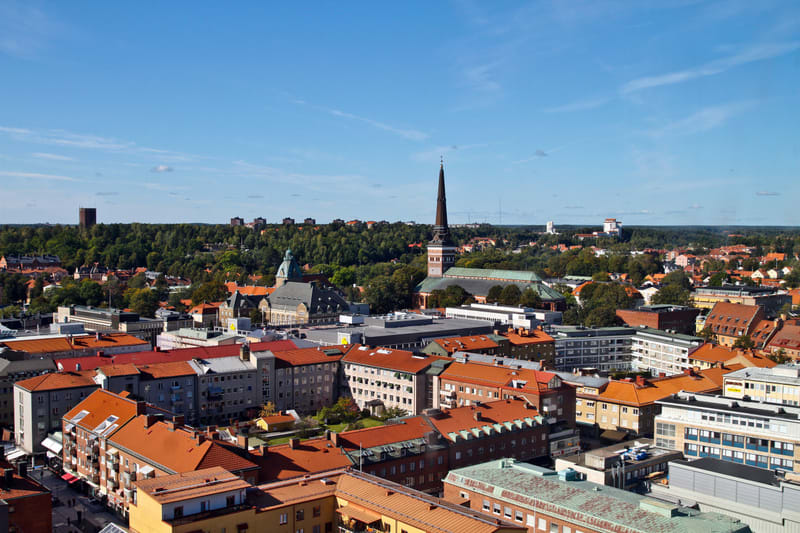 En översiktsbild av en stad sedd från ovan, med många hustak i rött och grått. En kyrka med en hög, spetsig spira syns i mitten, omgiven av träd och andra byggnader. I bakgrunden syns skog och en blå himmel med några moln.