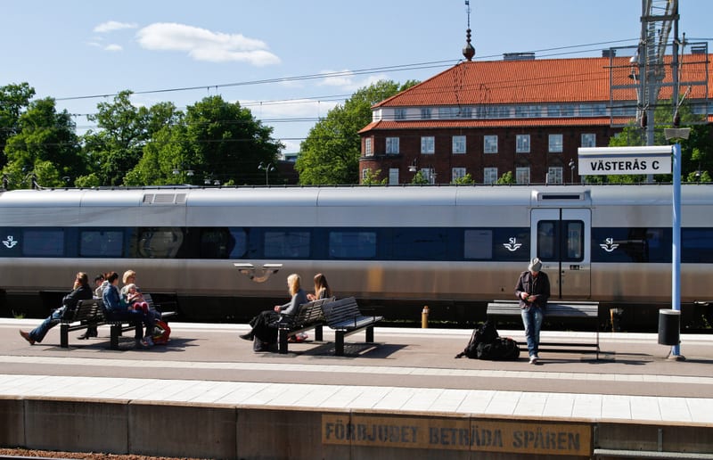 En bild som visar en tågperrong med ett SJ-tåg och flera personer som väntar. I bakgrunden syns träd och en byggnad med rött tak. En skylt med texten "Västerås C" är synlig.