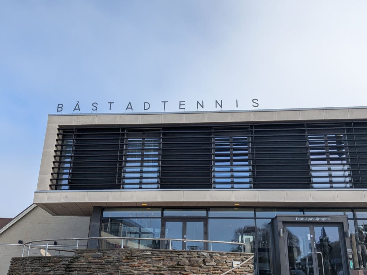 En modern byggnad med stora fönster och texten "BÅSTADTENNIS" på taket. Byggnaden har även en skylt med texten "Tennispaviljongen" vid entrén. Fasaden består av stenmaterial och glaspartier med svarta jalusier. En stentrappa med räcke syns i förgrund