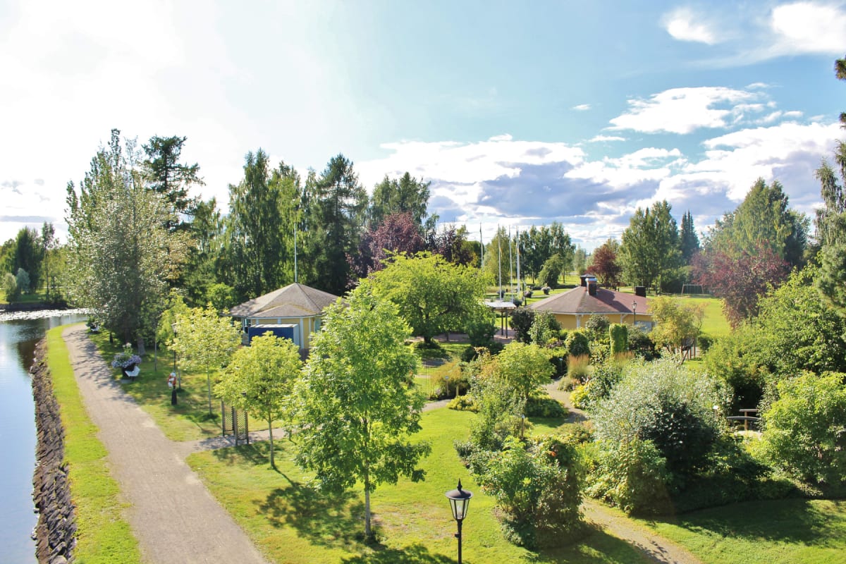 En översiktsbild över ett grönområde med byggnader, träd och buskar intill en vattenkanal under en klarblå himmel med moln. En grusväg löper längs kanalen och en gammaldags lyktstolpe syns i förgrunden.