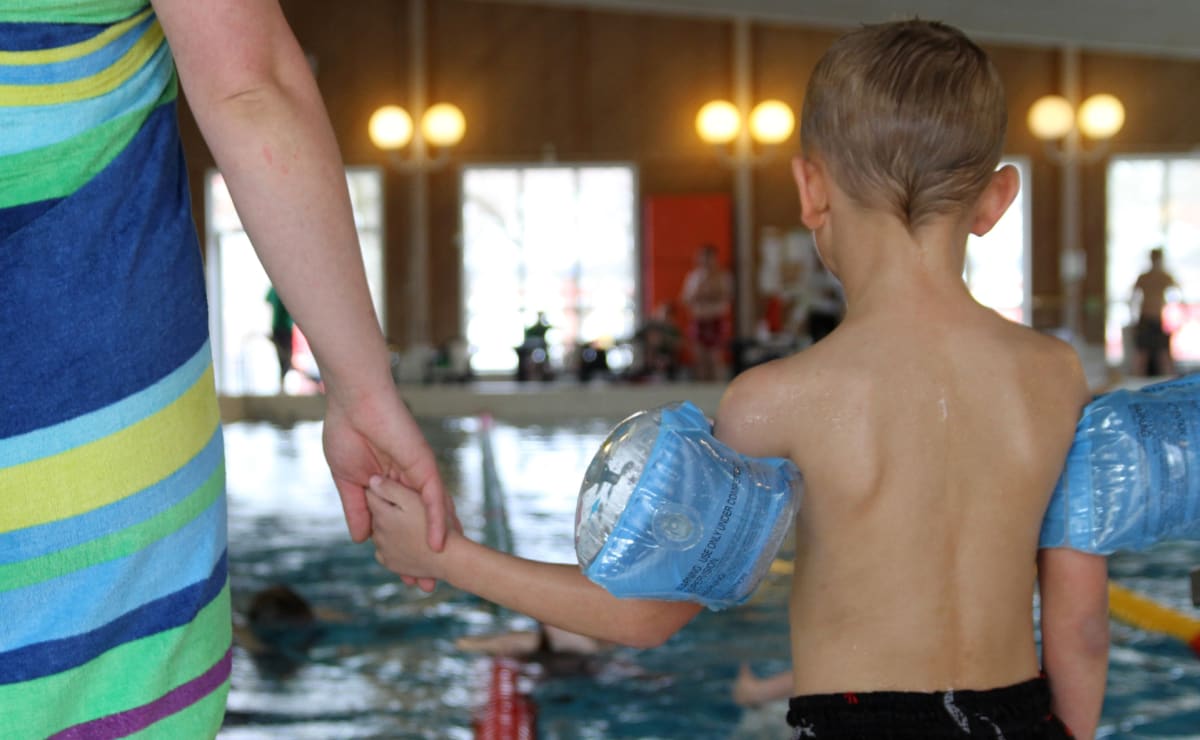 En person i en färgglad handduk som håller ett barns hand i en simbassäng. Barnet har blåa simdynor, är vänd bort från kameran och har vått hår. I bakgrunden syns andra personer och bassängen med vatten. Fönster och belysning reflekteras i vattnet.