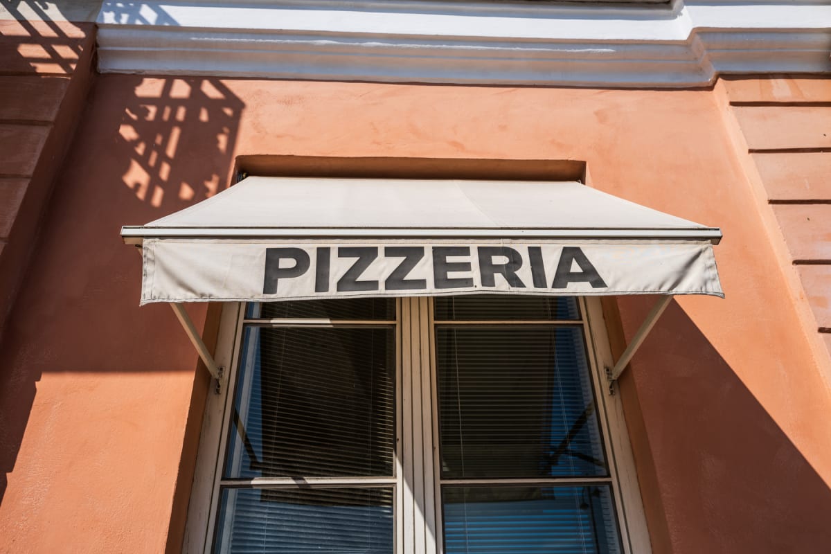 En beige markis med texten "PIZZERIA" i svarta bokstäver hänger över två fönster med persienner på en laxrosa byggnadsfasad. En vit list och gipsdekor syns över fönstret, och skuggor från något utanför faller på fasaden. Solen lyser på byggnaden.