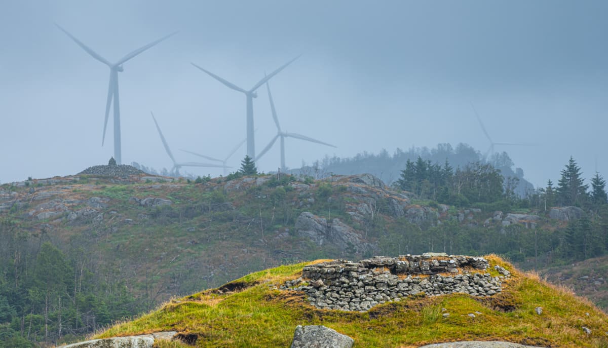 En bild visar ett landskap med kullar och gles vegetation under en disig himmel. Flera vindkraftverk är synliga i bakgrunden, delvis dolda av dis. I förgrunden finns en stenvägg på en grön kulle och en stencairn på en annan kulle längre bort.