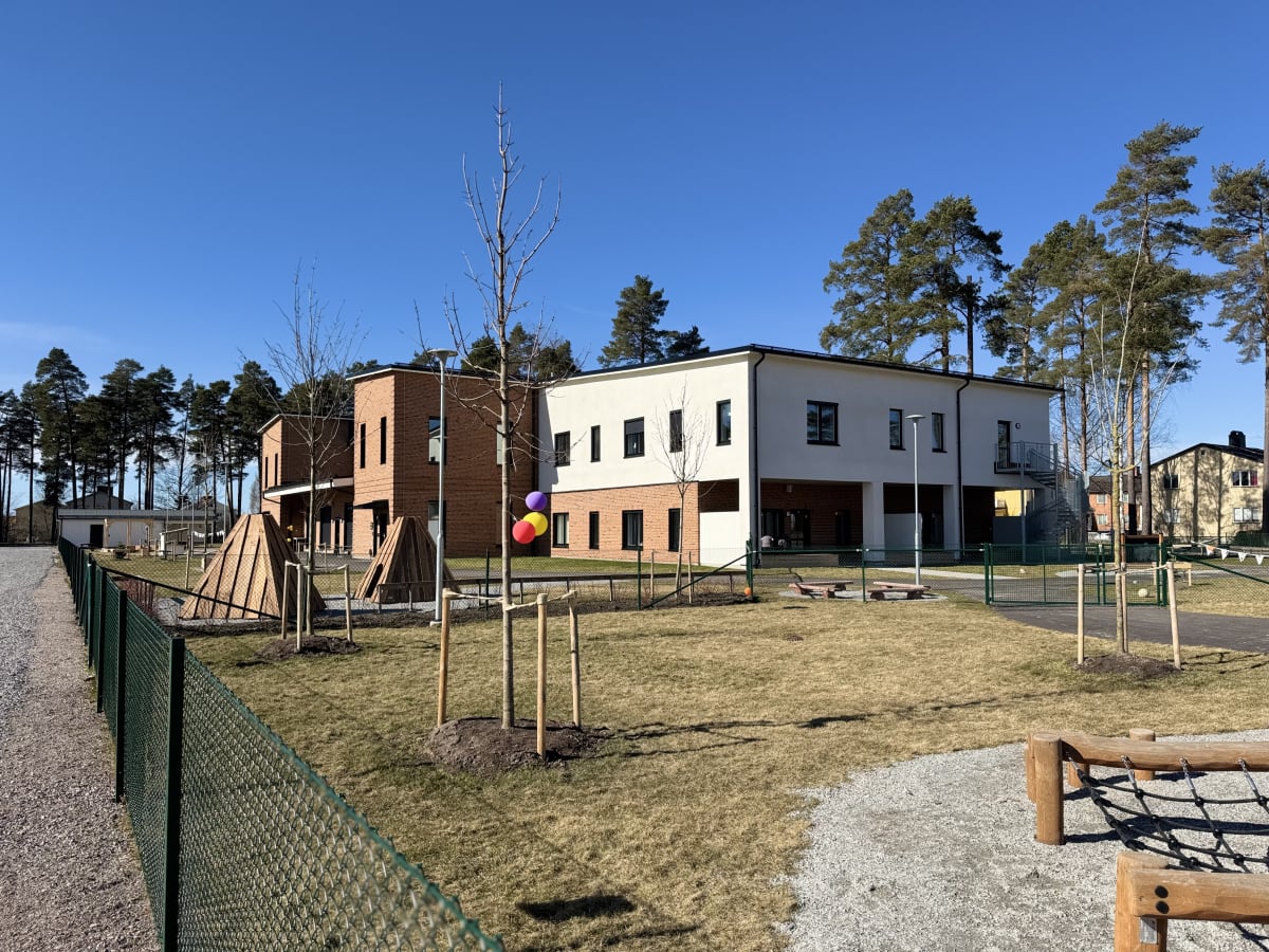 Bilden visar byggnader med beige och tegelröda fasader, samt ett utomhusområde med gräs, grus, träd och leksaker. En grön metallstaket löper genom bildens förgrund och mitt. Himlen är klarblå och inga moln syns.