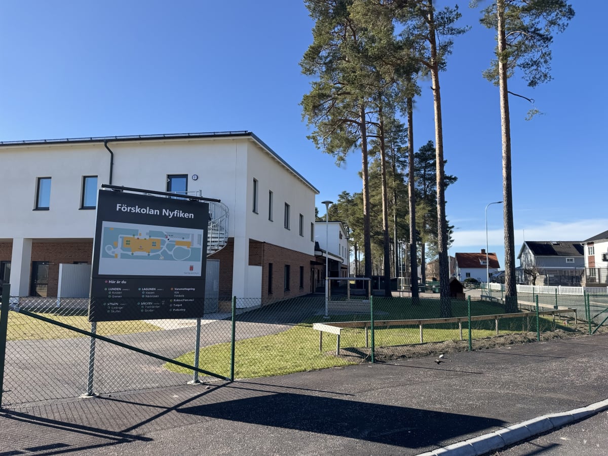 En modern byggnad med vit fasad och röda tegeldetaljer står under en klarblå himmel. Framför byggnaden finns en informationsskylt för "Förskolan Nyfiken" och i bakgrunden syns höga tallar och bostadshus. En grön metallstaket löper längs förgrunden.