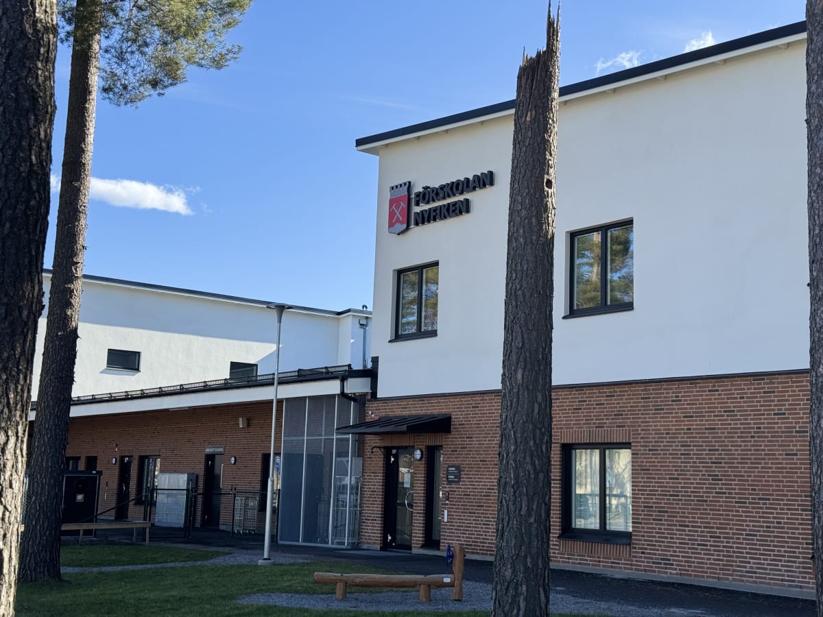 En modern byggnad med vit fasad och röd tegelbas, omgiven av tallar och gräs. På byggnaden finns en skylt med texten "FÖRSKOLAN NYFIKEN" och ett vapenliknande emblem. Himlen är klarblå med några vita moln.