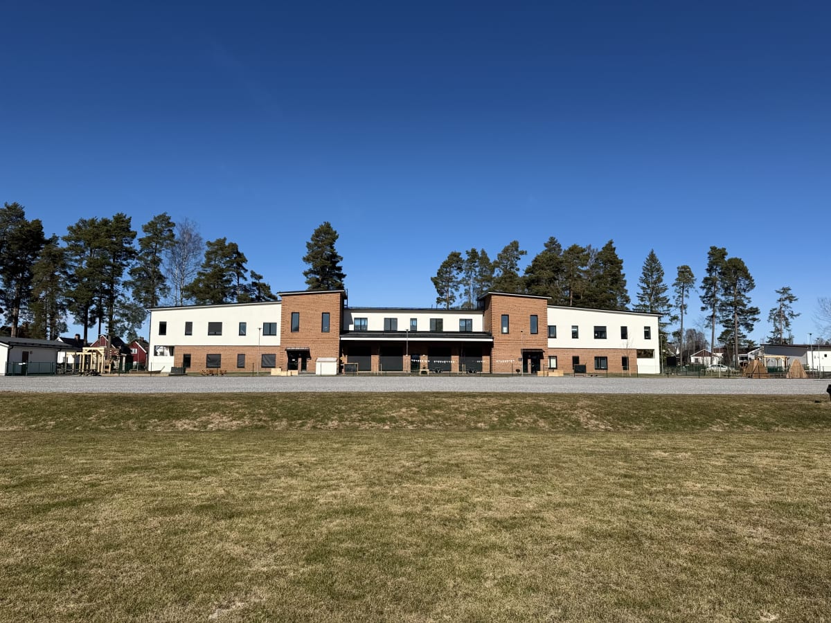 En modern byggnad med tegel och vit fasad under en klarblå himmel. Framför byggnaden finns ett grusområde och i förgrunden en gräsmatta. Bakom byggnaden syns tallar och några hus.