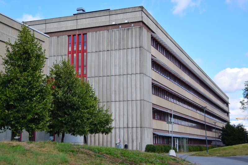En stor, flervåningsbyggnad med fasad av betong och rödbruna fönsterramar under en klarblå himmel. Delar av byggnaden har vertikala betongpaneler, andra delar har horisontella rader av fönster. Två träd med gröna löv syns i förgrunden.
