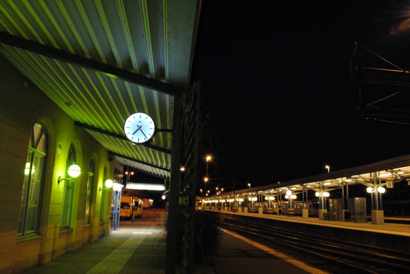En bild tagen nattetid som visar en upplyst tågstation eller perrong. Till vänster syns del av en byggnad med fönster och en klocka. Till höger sträcker sig perronger och tågspår under mörk himmel, belysta av stationens lampor.