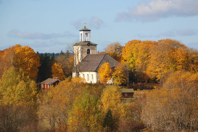 En kyrka belägen på en kulle omgiven av träd med löv i gula och orangea höstfärger, under en blå himmel med vita moln. Bredvid kyrkan finns en röd byggnad och en till röd mindre byggnad syns längre bort. Ett staket syns runt kyrkogården.