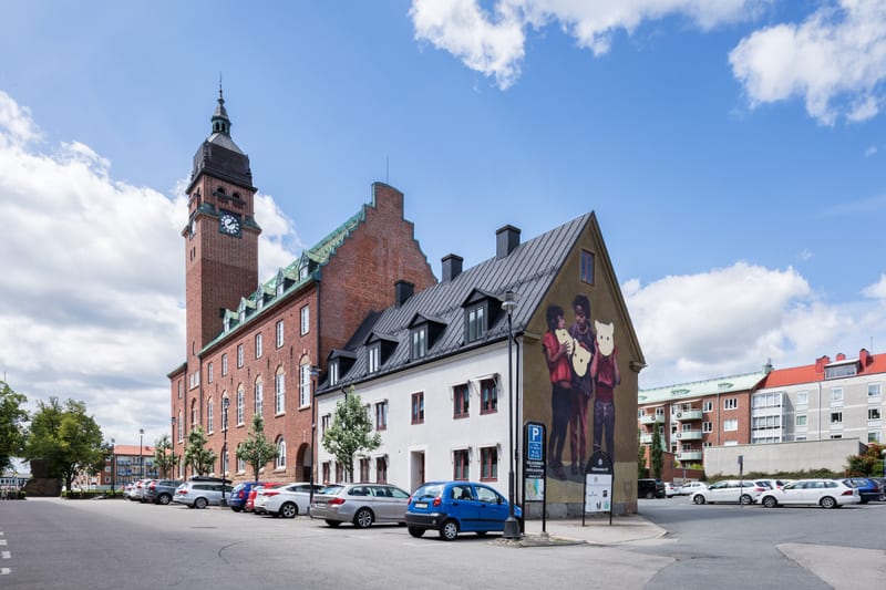 En exteriörbild som visar en gatuparkering med flera bilar framför en stor tegelbyggnad med klocktorn. Till höger om parkeringen står ett gult hus med en färgglad väggmålning som avbildar tre figurer. Byggnaden till höger om det gula huset är ett fle