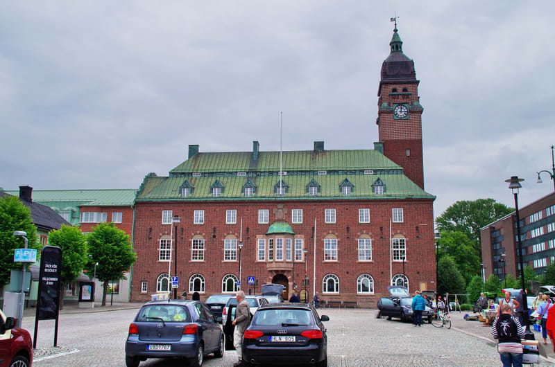 En stor, röd tegelbyggnad med grönt tak och ett klocktorn dominerar bilden. Framför byggnaden finns en kullerstenstorg med parkerade bilar och människor. På torget syns även en gatumarknad med stånd och besökare.