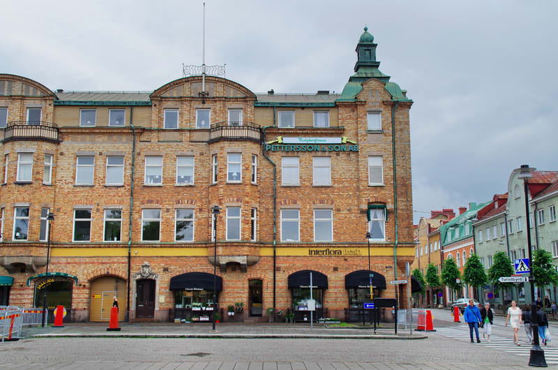 En stor, tegelröd byggnad i flera våningar med vita fönster, balkonger och ett grönt torn. På fasaden finns skyltar för "PETTERSSON & SON AB" och "interflora fresh". Gator syns med trafikkoner, gatuskyltar och en zebraövergång där flera personer går.