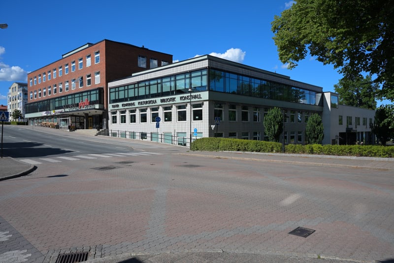 En väg med korsningar och två större byggnader på en solig dag. Den vänstra byggnaden är tegelröd med skyltar som nämner "Kulturhuset" och "Statt". Den högra byggnaden är ljusgrå med stora fönster och texten "TEATER KONFERENS KULTURSKOLA BIBLIOTEK KO