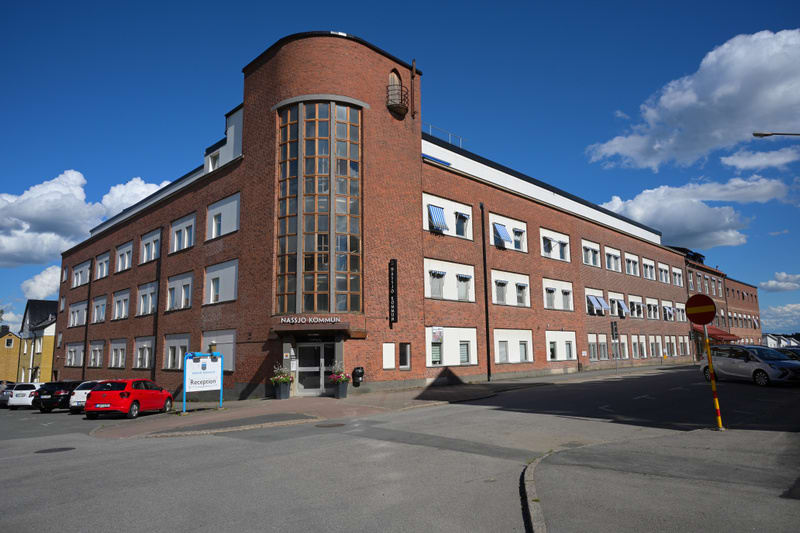 En stor röd tegelbyggnad under en blå himmel med vita moln dominerar bilden. Byggnaden har många fönster och en rundad sektion med stora glasytor på ena hörnet. En skylt med texten ”Nässjö Kommun” är synlig på byggnaden, samt en skylt för ”Reception”