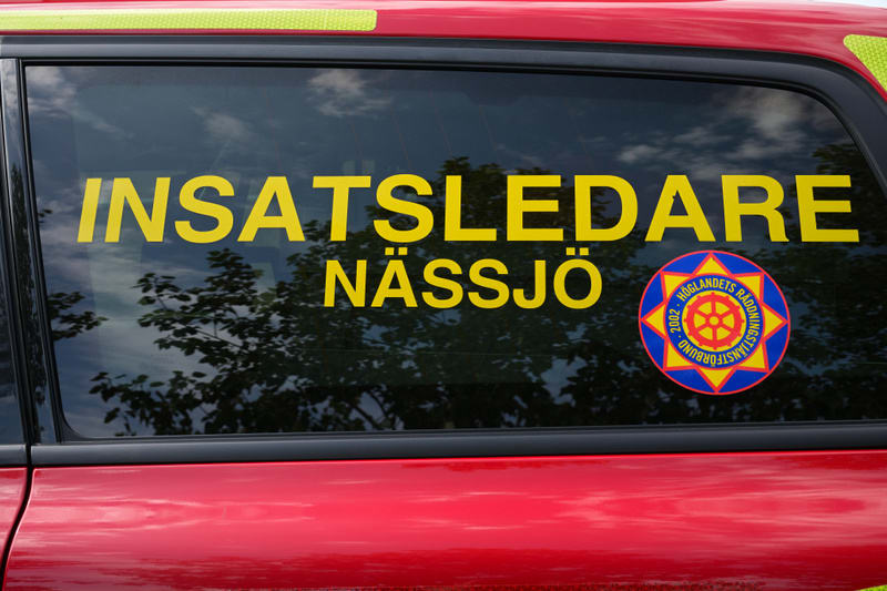 En bild visar en detalj av sidan på ett rött fordon med mörka fönster. På fönstret sitter gula textdekaler som bildar orden "INSATSLEDARE NÄSSJÖ" och en rund dekal med texten "HÖGLANDETS RÄDDNINGSNÄNSTFÖRBUND 2002". Träd och moln speglas i fönstret.