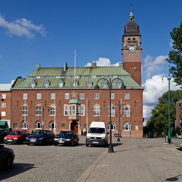 En stor röd tegelbyggnad med grön tak och ett klocktorn dominerar bilden under en blå sommarhimmel med några moln. Framför byggnaden finns ett bilparkering med flera personbilar och en vit skåpbil. Till höger syns gatlyktor och träd.