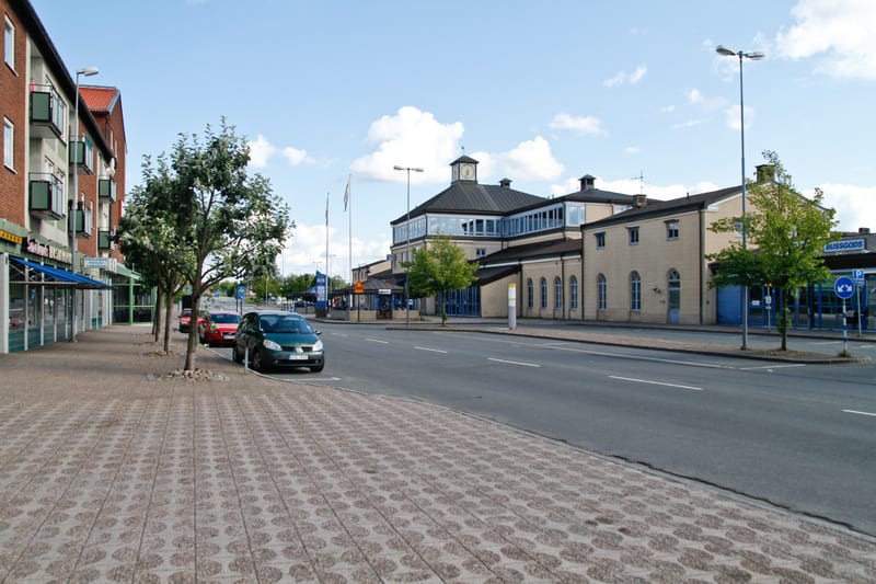 En gata med parkerade bilar och träd på vänster sida. Till vänster syns en röd tegelfastighet med balkonger och butikslokaler i markplan. Till höger syns en större ljusgul byggnad med klocktorn och en skylt "BUSSGODS" samt en parkeringsskylt.
