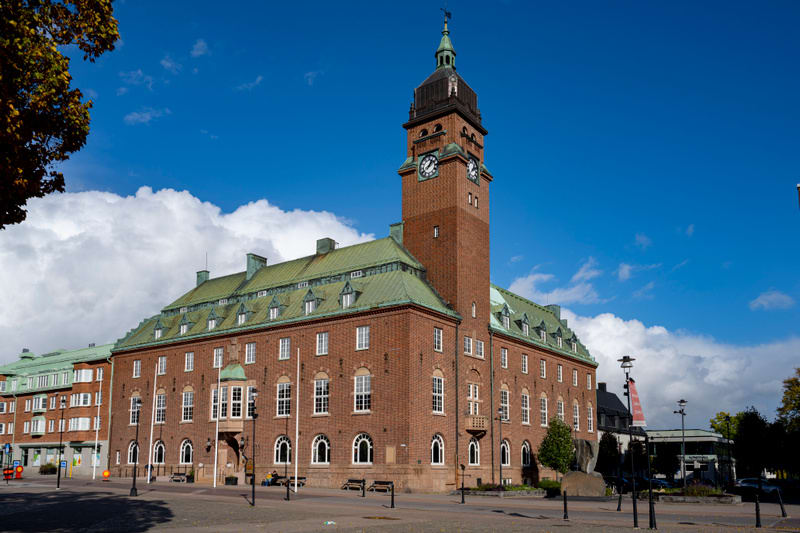 En stor, röd tegelbyggnad med ett högt klocktorn och ett grönt tak dominerar bilden. Byggnaden har många fönster och balkonger. I förgrunden syns en öppen plats med gatlyktor, några bänkar samt träd och buskar. Himlen är blå med vita moln.