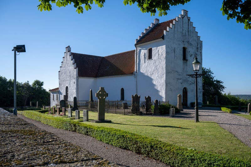 En vit kyrka med rött tak och trappstegsgavlar står under en klarblå himmel. Runt kyrkan finns en kyrkogård med gravstenar, en gräsmatta, buskar och träd. En grusgång och en gatlykta syns också.