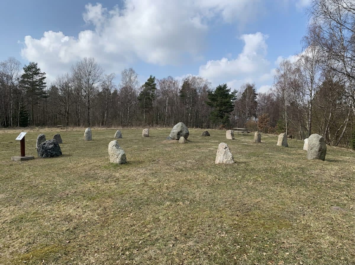 En utomhusbild tagen under dagtid visar en stencirkel på ett gräsfält. Cirka 15 synliga stenar av olika storlek är placerade i gräset. I bakgrunden syns skog med, dels barrträd och dels lövträd som saknar löv. En informationsskylt står till vänster i