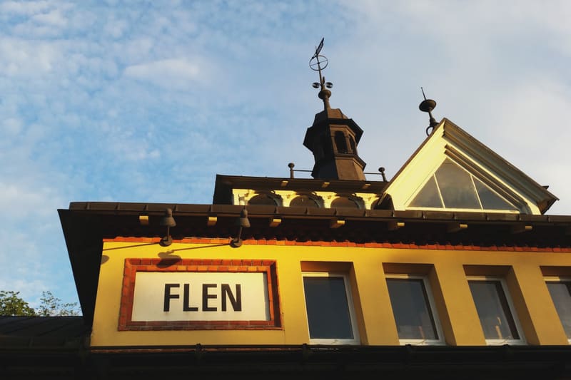 Bilden visar delar av en gul byggnad med tak och en skylt där "FLEN" står skrivet i svarta bokstäver mot vit bakgrund. Ovanpå taket syns en väderflöjel och takfönster. Byggnaden är belyst av solljus från höger, vilket skapar skuggor. Bakom byggnaden