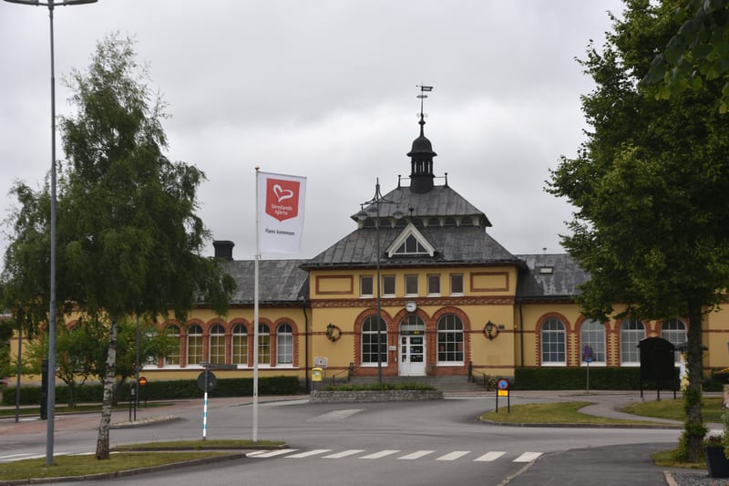En gul byggnad med grått tak och en tornspira med vindflöjel står i mitten av bilden. Framför byggnaden finns en väg med övergångsställen och en flaggstång med en vit flagga. Runtomkring växer gröna träd och buskar, och himlen är molnig och grå.