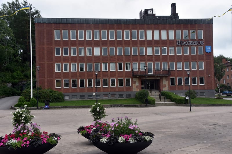 En fasadvy av en röd tegelsbyggnad med skylten "STADSHUS" ovanför ingången. Framför byggnaden finns ett öppet torg med större blomkärl fyllda med växter i olika färger, samt en flaggstång med en vajande svensk flagga. I bakgrunden syns skog och en ku