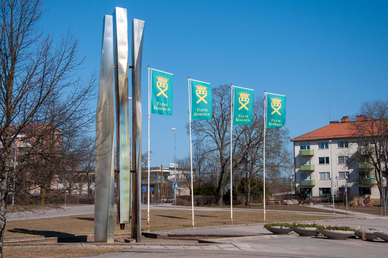 En utomhusbild som visar en modern metallskulptur och fyra turkosa flaggor med texten "FLENS KOMMUN" och ett guldfärgat vapen under en klarblå himmel. I bakgrunden syns kala träd, en ljus byggnad med orange taktegel och gräsytor.