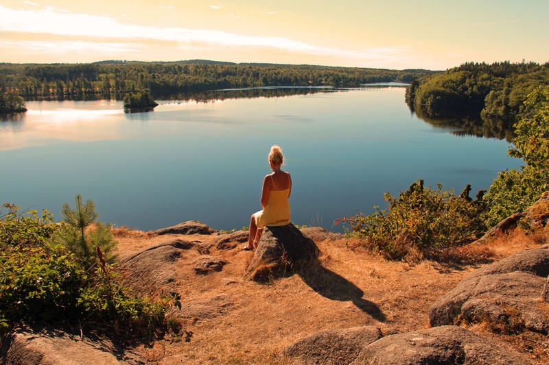 En kvinna i gul klänning sitter vänd bort från kameran på en klippa och tittar ut över en sjö omgiven av skog. Solen står lågt på himlen och kastar långa skuggor på marken. Vidsträckt utsikt över ett vattenlandskap med grönskande skog.