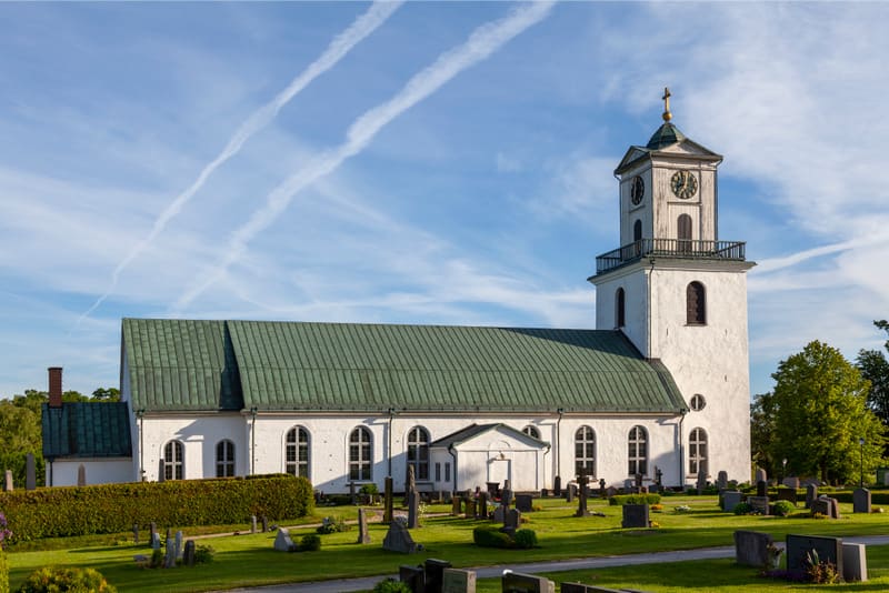 En vit kyrka med grönt tak och en klocka i tornet står under en blå himmel med flygplansstrimmor. Runt kyrkan finns en grön kyrkogård med gravstenar och träd, och en grön häck löper längs en del av kyrkobyggnaden.