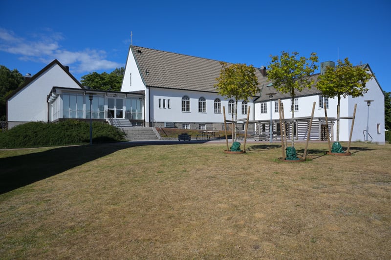 En stor vit byggnad med ett tegeltak och flera fönster under en klarblå himmel. Framför byggnaden finns en klippt gräsmatta som är delvis torr. Tre unga träd med löv är planterade på gräsmattan, var och en stödd av stolpar och med en grön påse vid ba
