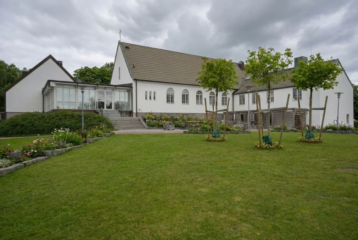 En stor vit byggnad med ett tegeltak och många fönster. Framför byggnaden finns en klippt gräsmatta, blomsterrabatter och tre små träd med stödpinnar. Himmelen är mulen.