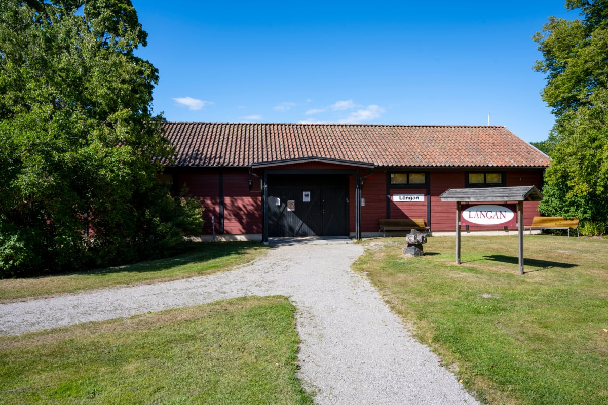 En röd byggnad med tegelpannor på taket. Framför byggnaden finns en grusgång som förgrenar sig och gräsytor. Stora gröna träd ramar in byggnaden på sidorna. En skylt med texten "LÅNGAN" står till höger om grusgången.