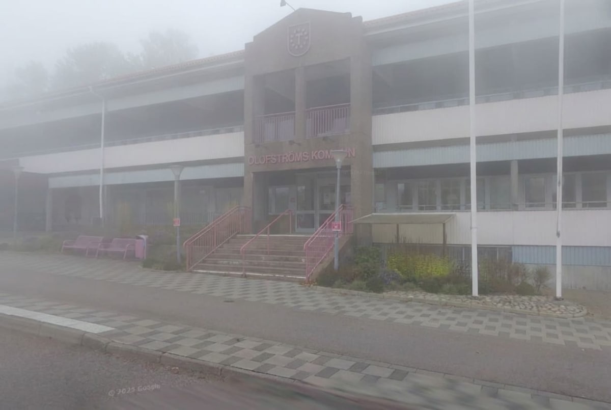 En bild av Olofströms kommunhus i dimma eller tjockt dis. Byggnaden har en stenfasad med ett kommunvapen ovanför entrén, röda trappor och räcken, samt vita långsträckta fönsterpartier. Markbeläggningen framför byggnaden består av en kombination av gr