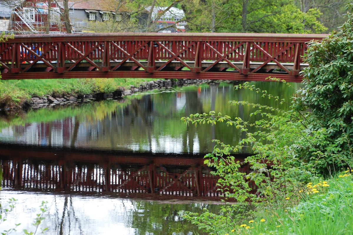 En röd träbro sträcker sig över en vattenfylld kanal med gröna stränder. Vårliknande växtlighet med ljusgröna blad syns i förgrunden och spegelbilder av bron och omgivningen reflekteras i vattnet. I bakgrunden syns delar av byggnader och träd.