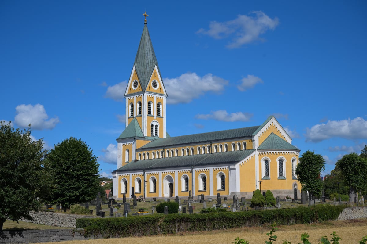 En gul kyrka med ett högt, grått spetsigt tak och ett mindre, grönt tak. Framför kyrkan finns en gräsmatta med gravstenar och ett buskage. I förgrunden syns torkat gräs och stenmurar, samt en del gröna träd mot en klarblå himmel med spridda moln.