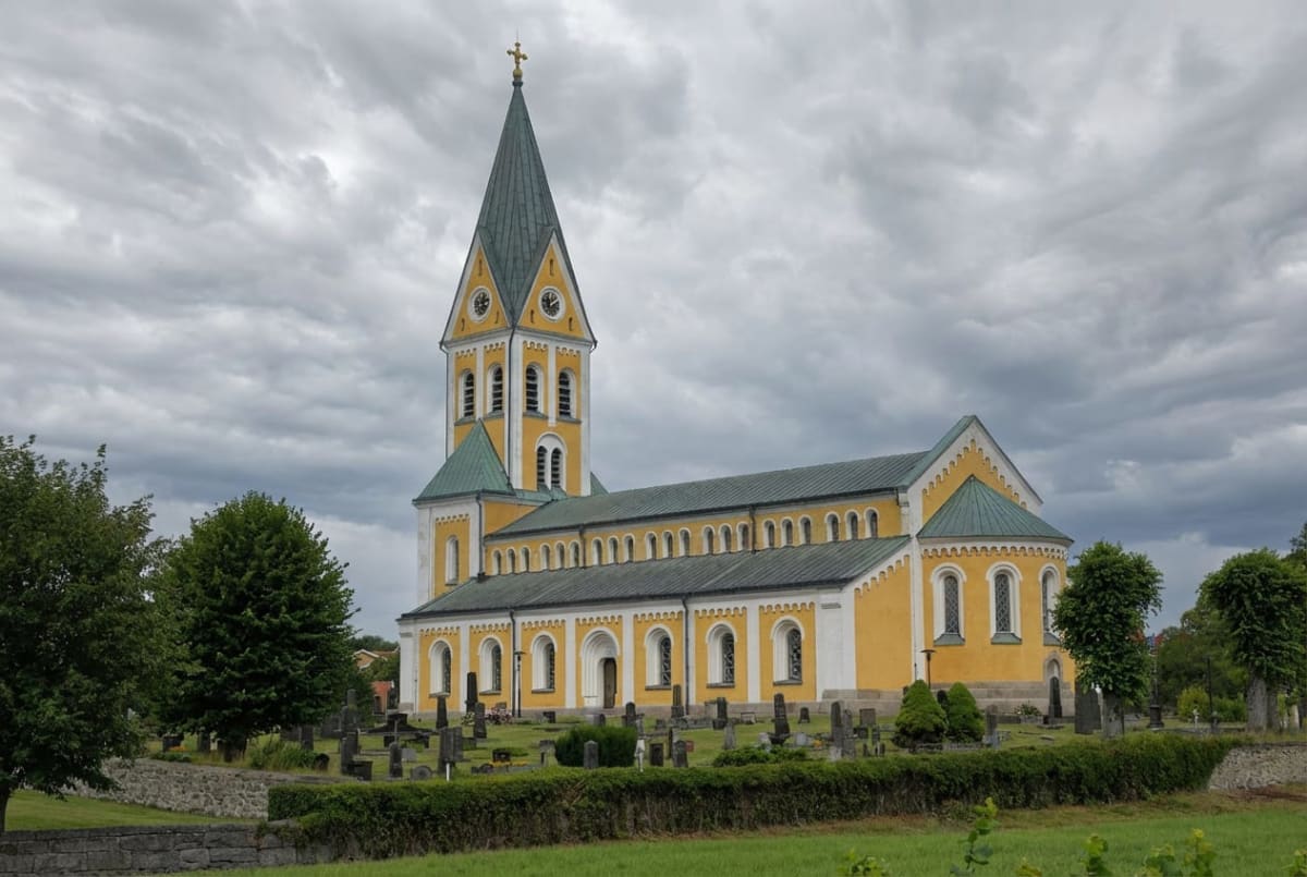 En gul kyrka med ett spetsigt grönt tak och ett klocktorn, omgiven av en kyrkogård med gravstenar och träd under en molnig himmel. Kyrkan har många fönster med rundade valv. Framför kyrkan syns gräs och en stenmur.