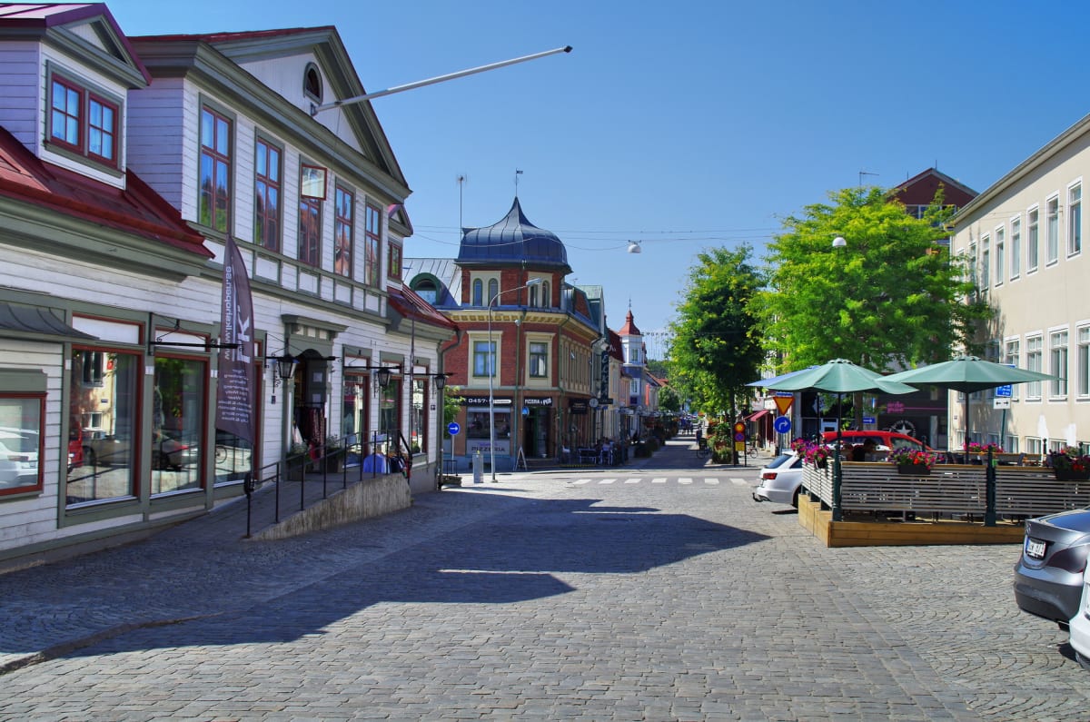 En gata med gatsten belyst av solsken kantad av flervåningshus i olika stilar med butiker och restauranger på bottenvåning. Träd med gröna löv och uteserveringar med parasoller syns längs gatan. Flera bilar är parkerade vid sidan av gatan.