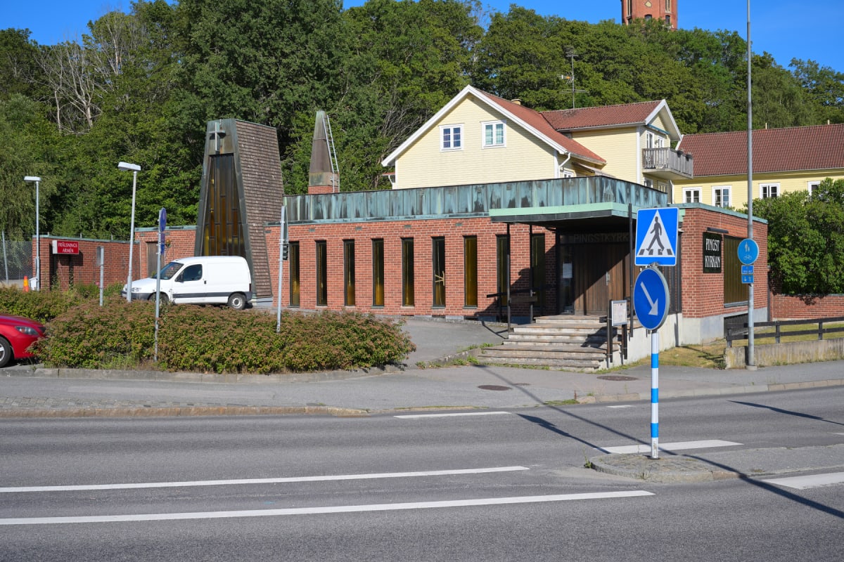 En bild visar en gata i förgrunden och byggnader i bakgrunden. Ett vitt servicefordon står parkerat på en gräsplätt vid en tegelbyggnad. Två trafikskyltar, en för övergångsställe och en för påbjuden körriktning, syns i närheten av en byggnad med skyl
